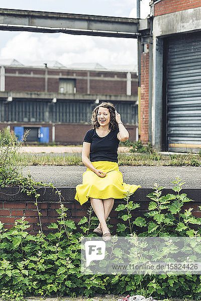 Glückliche Frau sitzt auf einer mit Pflanzen bedeckten Wand in einem alten Industriegebiet
