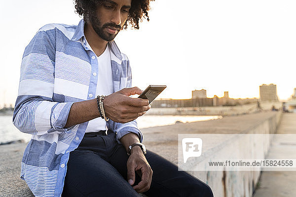 Junger Mann sitzt bei Sonnenuntergang mit einem Smartphone an der Kaimauer