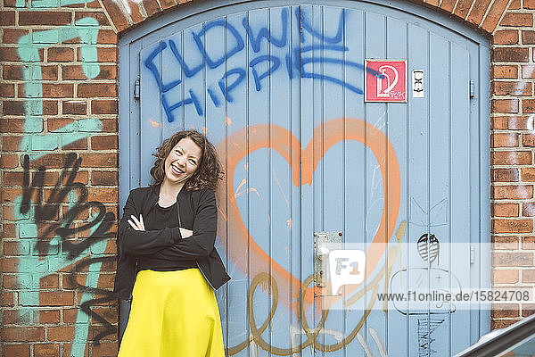 Porträt einer glücklichen brünetten Frau  die vor einer Ziegelmauer mit einem alten Tor und Graffiti steht