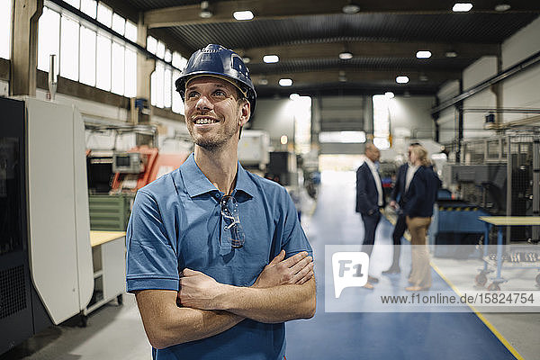 Portrait of a confident worker in a factory wearing hard hat with business people in background