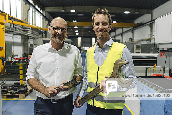 Portrait of businessman and man in reflective vest with large wrenches in a factory