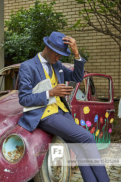 Junger Geschäftsmann in altmodischem Anzug und Hut lehnt an Oldtimer und überprüft sein Telefon