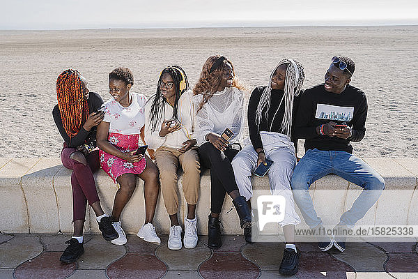 Glückliche Teenager-Freundinnen und junger Mann sitzen mit Smartphones an einer Wand am Strand