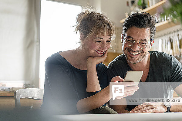 Happy couple sitting at table in kitchen using smartphone