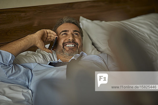 Laughing businessman lying on the bed in hotel room talking on the phone