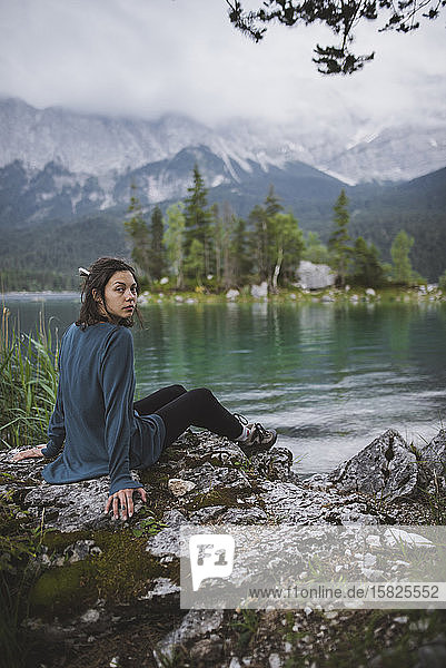 Germany  Bavaria  Eibsee  Portrait of young woman sitting on rock at shore of Eibsee lake in Bavarian Alps