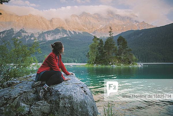 Deutschland  Bayern  Eibsee  Junge Frau sitzt auf einem Felsen und blickt auf eine malerische Landschaft am Eibsee in den bayerischen Alpen