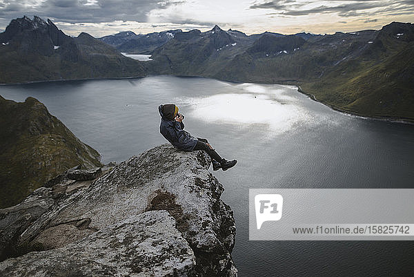 Norwegen  Senja  Mann fotografiert sitzend am Rande einer steilen Klippe auf dem Gipfel des Berges Segla