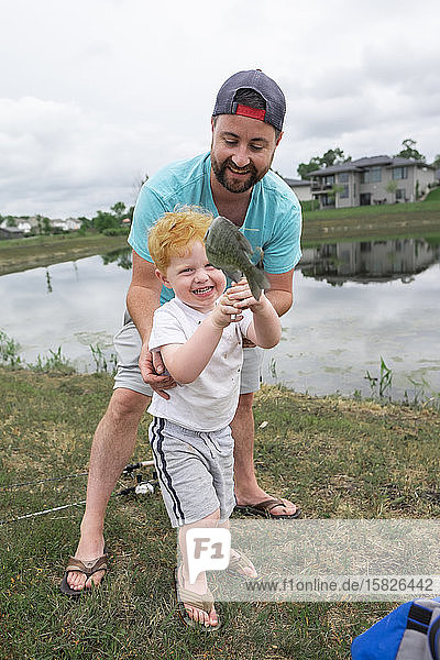 Smiling Father helps toddler son hold up a fish he got at a pond