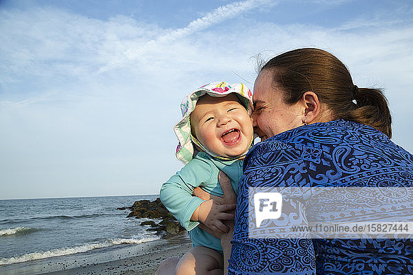 Mutter umarmt glücklich lächelndes Mädchen am Strand sitzend
