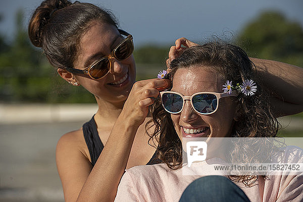 Zwei Frauen draußen  die lachen und Blumen ins Haar stecken.