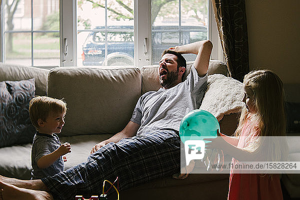 A tired father lies on the couch while his children play around him.