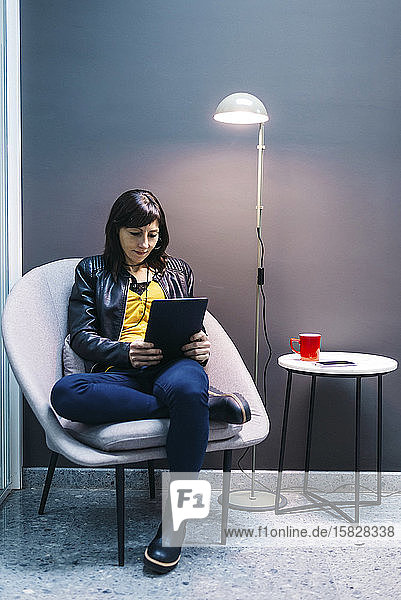 Frau zu Hause  die auf einem modernen Stuhl sitzt und einen Tablet-Computer benutzt