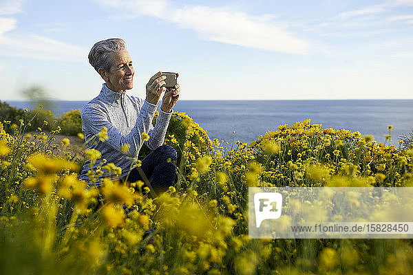 Lächelnde ältere Frau fotografiert mit einem Smartphone  während sie am Meer bei Pflanzen gegen den Himmel kauert