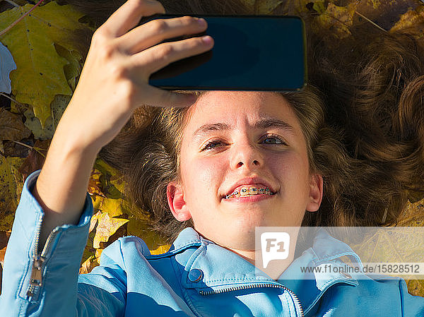 Teenager-Mädchen mit brünetten Haaren  das auf dem Boden liegt und mit gelben Blättern bedeckt ist  während sie mit ihrem Handy ein Foto für sich selbst macht. Das Mädchen lächelt in die Kamera. Horizontales Foto