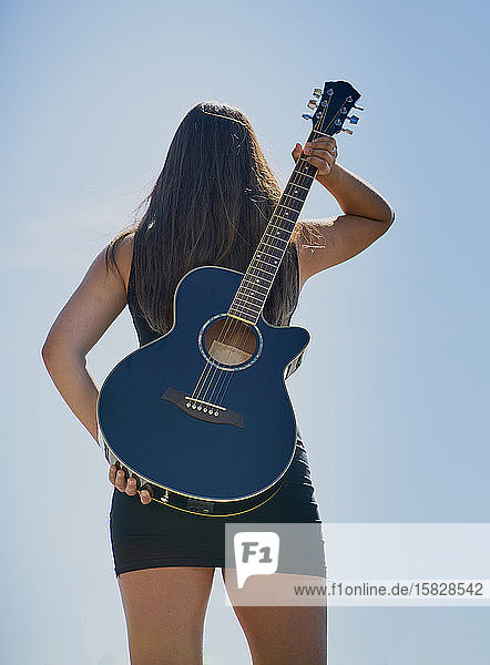 Eine jugendliche Gitarristin hält sich eine Akustikgitarre auf dem Rücken vor einen blauen Himmel