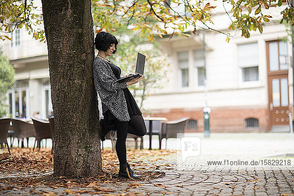 junge Frau mit Laptop im Freien  die in der Nähe eines Baumes arbeitet
