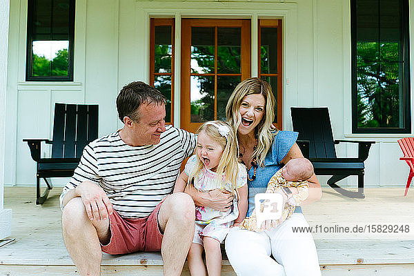 Eine vierköpfige Familie lacht zusammen  während sie auf einer modernen Veranda sitzt