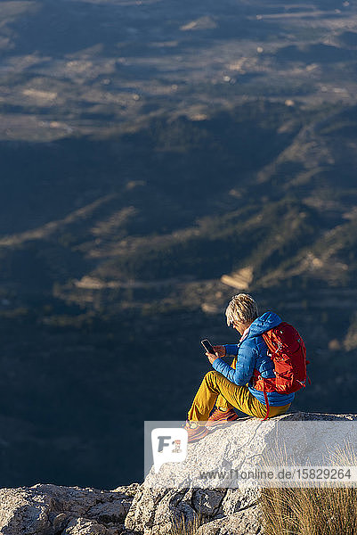 Junge Frau  die auf Felsen auf einem Berggipfel mit einem Mobiltelefon steht.