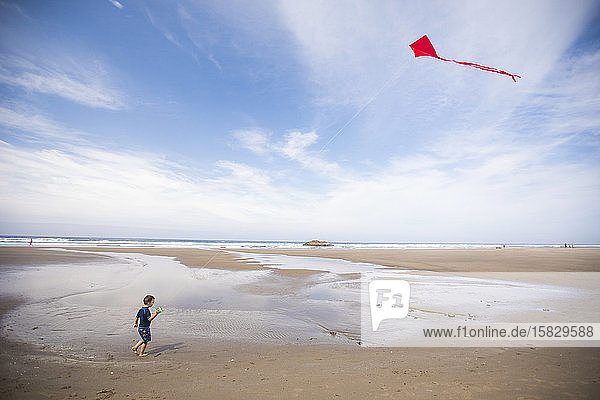 Toddler boy flying red kite at beach.