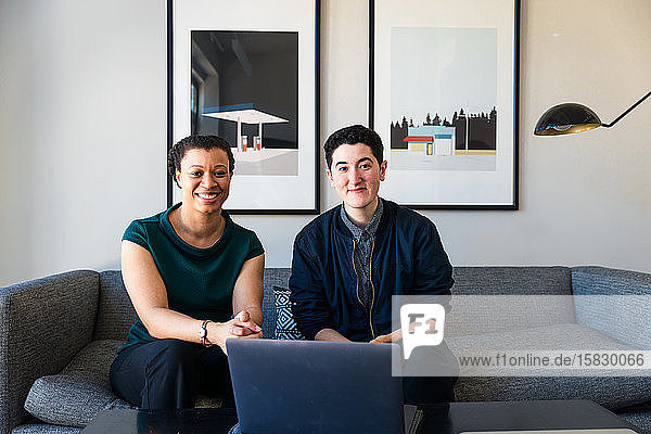 Portrait of smiling business colleagues sitting with laptop at table against wall in office