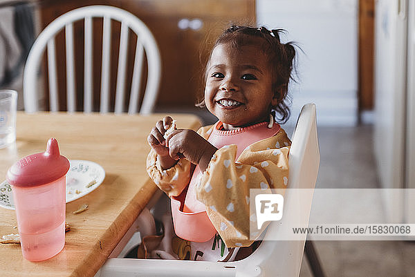Lächelndes 2 Jahre altes gemischtrassiges Mädchen mit Lätzchen im Hochstuhl in der Küche