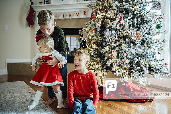 Brothers holding baby sister in front of Christmas tree and fireplace