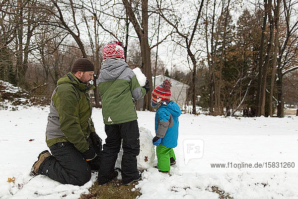 Father kneels on ground next to two sons while making a snowman