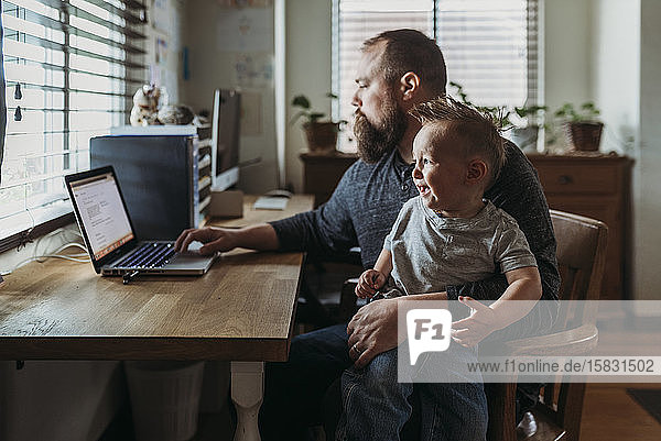 Vater arbeitet von zu Hause aus mit einem einjährigen Jungen  der auf dem Schoß weint