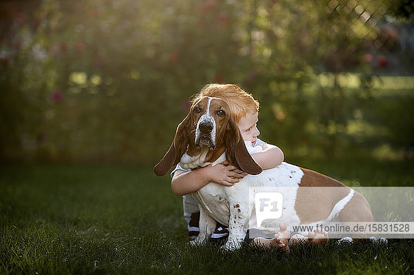 Toddler boy sitting and hugging his hound dog in the backyard grass