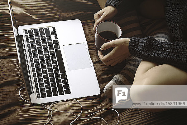 Junges Mädchen im Winter stilvolle Hähne sitzen auf dem Bett mit einer Tasse Kaffee und schauen sich etwas auf dem Laptop an. Winter  gemütlich  Kleidung und Lifestyle-Konzept.
