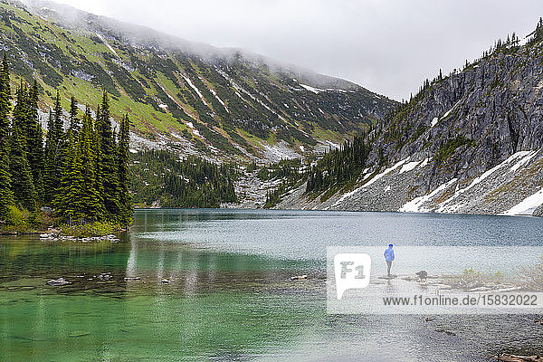 Eine Frau hängt mit ihrem Hund am Ufer eines Alpensees herum und nimmt an einem verregneten Sommertag die Aussicht in den Coast Mountains um Pemberton  British Columbia  auf.