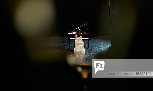a girl dressed in white with her back to the audience plays a black grand piano on a stage.