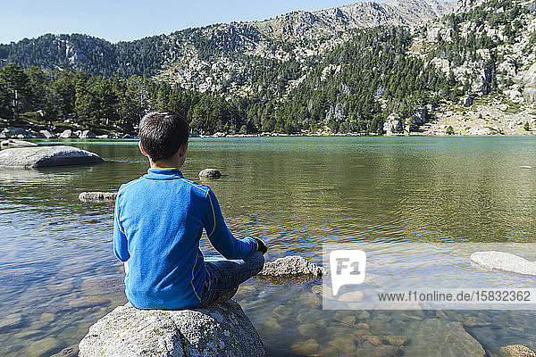 Rear view of a boy sitting on rock on a mountain lake looking away