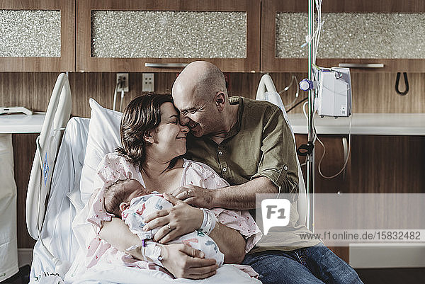 Porträt eines Vaters  der ein Neugeborenes und seine Frau im Krankenhausbett umarmt