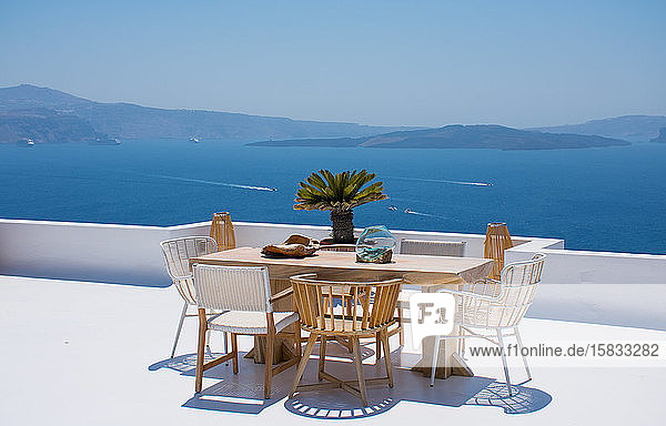 Möbel  bestehend aus einem Tisch und einigen Stühlen  auf einer weißen Terrasse eines Hauses in Santorin in Griechenland  wo man das Essen genießen kann  während man eine romantische Meereslandschaft bis hin zum blauen Ägäischen Meer sieht. Horizontal ph