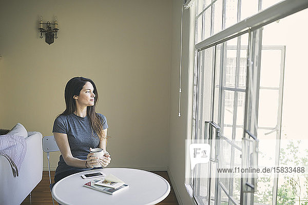 Nachdenkliche junge Frau trinkt Kaffee  während sie zu Hause durch das Fenster im Wohnzimmer schaut