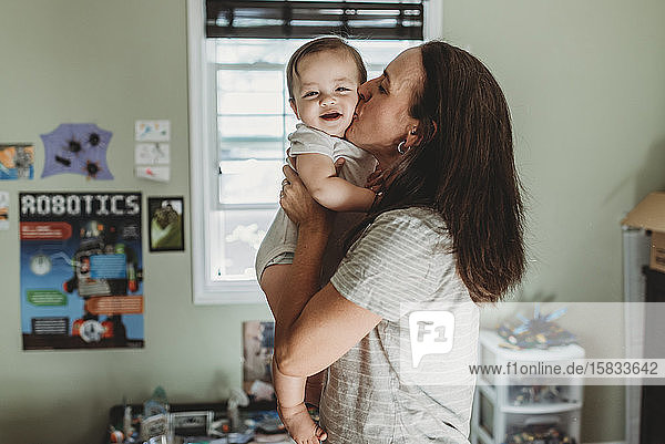 Loving mom kissing smiling baby in front of window in toy room