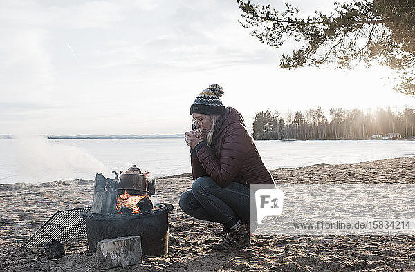 woman warming up sat next to a camp fire on an empty beach in Sweden