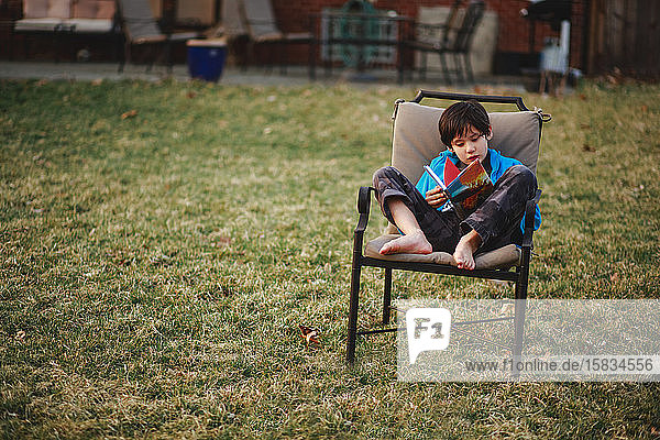 Ein kleines Kind sitzt barfuss auf einem Stuhl im Hof und liest im Frühling ein Buch