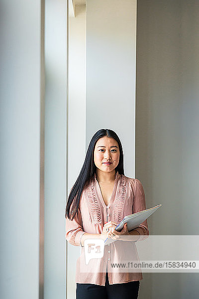 Porträt einer selbstbewussten Geschäftsfrau mit Akte  die im Büro an der Wand steht