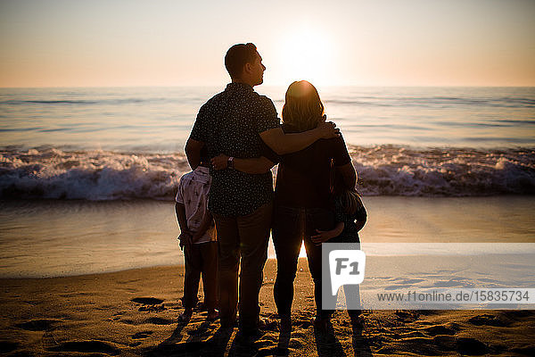 Vierköpfige Familie steht am Strand und schaut auf den Ozean bei Sonnenuntergang