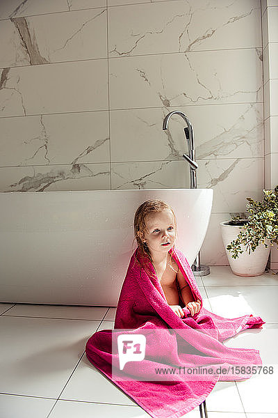 kleines Mädchen  das in einem rosa Handtuch auf dem Boden in der Badewanne sitzt