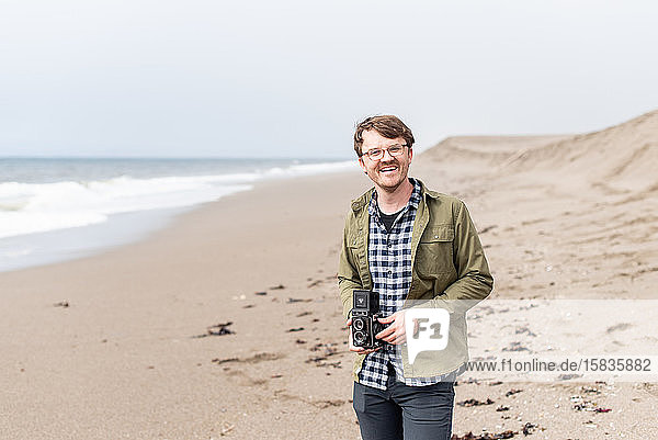 Porträt eines jungen Mannes  der lacht  während er eine Filmkamera am Strand hält