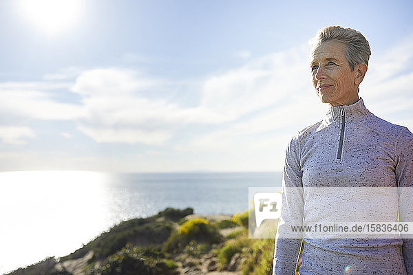 Ältere Frau schaut weg  während sie an einem sonnigen Tag auf einer Klippe am Meer gegen den Himmel steht