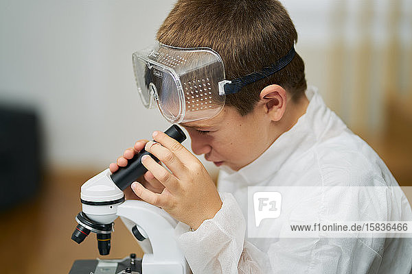 Junge in weißer Robe und Schutzbrille beobachtet durch ein Mikroskop in seinem Haus