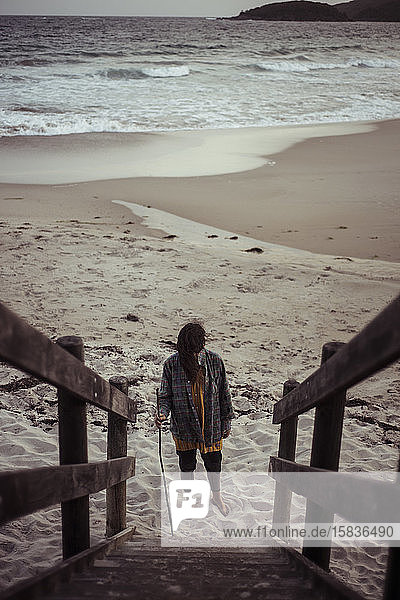 androgynes Mädchen steht am Fuße der Strandtreppe  die zum launischen Ozean führt