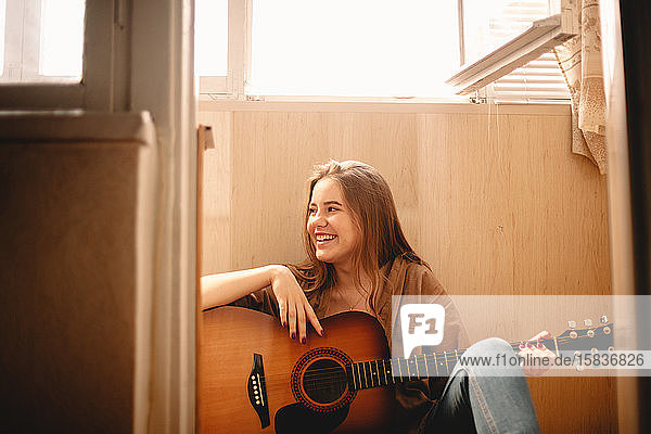 Fröhliche junge Frau hält Gitarre  während sie auf dem Balkonboden sitzt
