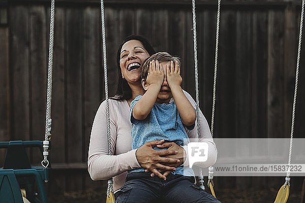 Young boy on swing with laughing mom covers his eeditorial