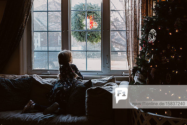 Ein kleines Mädchen sitzt neben einem Weihnachtsbaum und schaut aus einem Fenster.